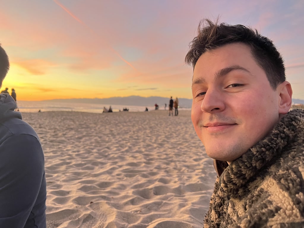 A person with short, styled hair and a serene expression is posing for a close-up photo, with a sunset and a beach setting in the background. The warm glow of the sunset softly illuminates the person's face, and the sky is painted in hues of orange, pink, and blue. Several people can be seen in the distance, some sitting on the sand. The sandy beach is textured and appears untouched around the individual, adding to the tranquil evening atmosphere.
